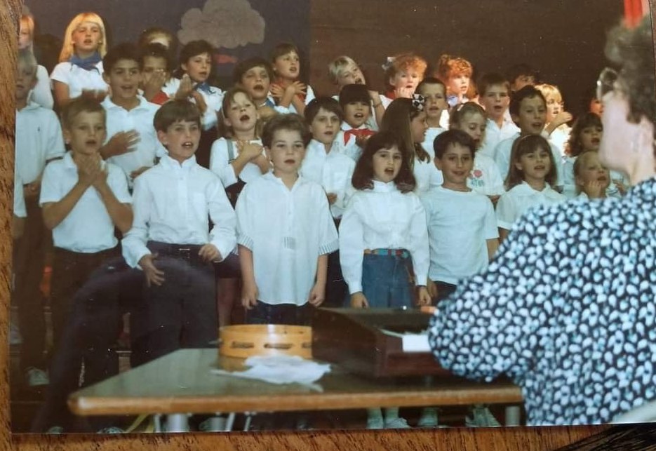 A school choir on stage, children in white shirts mid-song, their teacher at the keyboard
