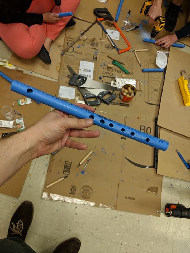 A student's hand holding a blue PVC flute with drilled finger holes over a workshop floor of tools and cardboard