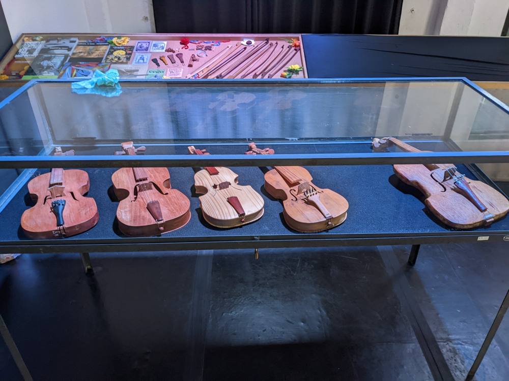 Five rabecas in a glass case, with a case of bows and album covers behind