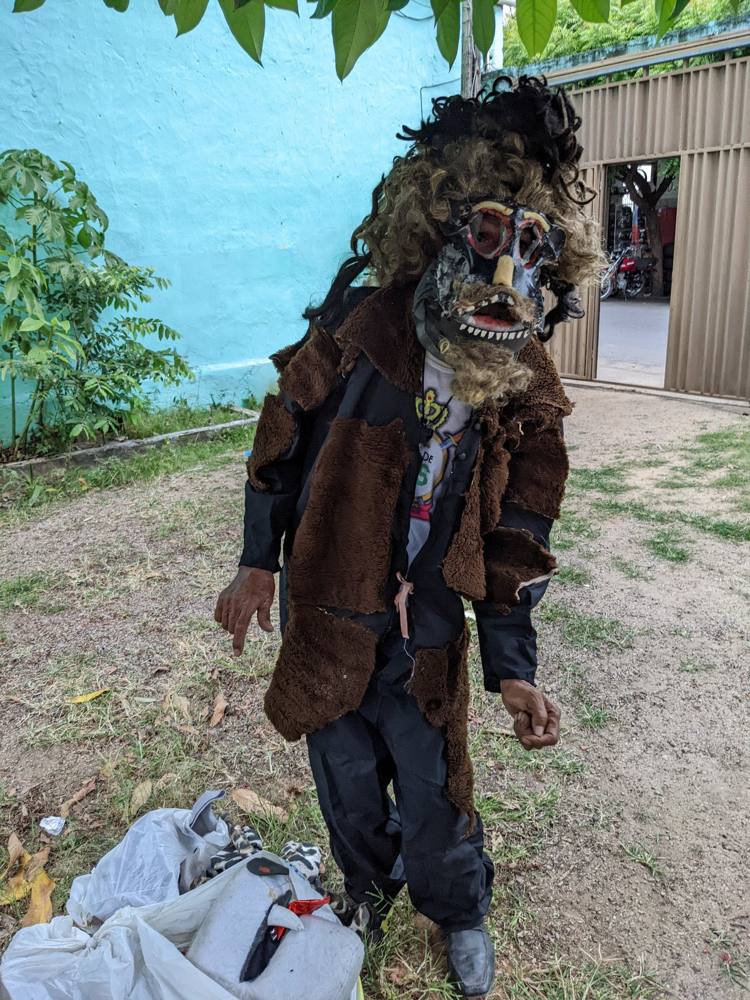 A performer in a folk-drama costume: a fierce mask with teeth, a shaggy wig, and a patched brown coat