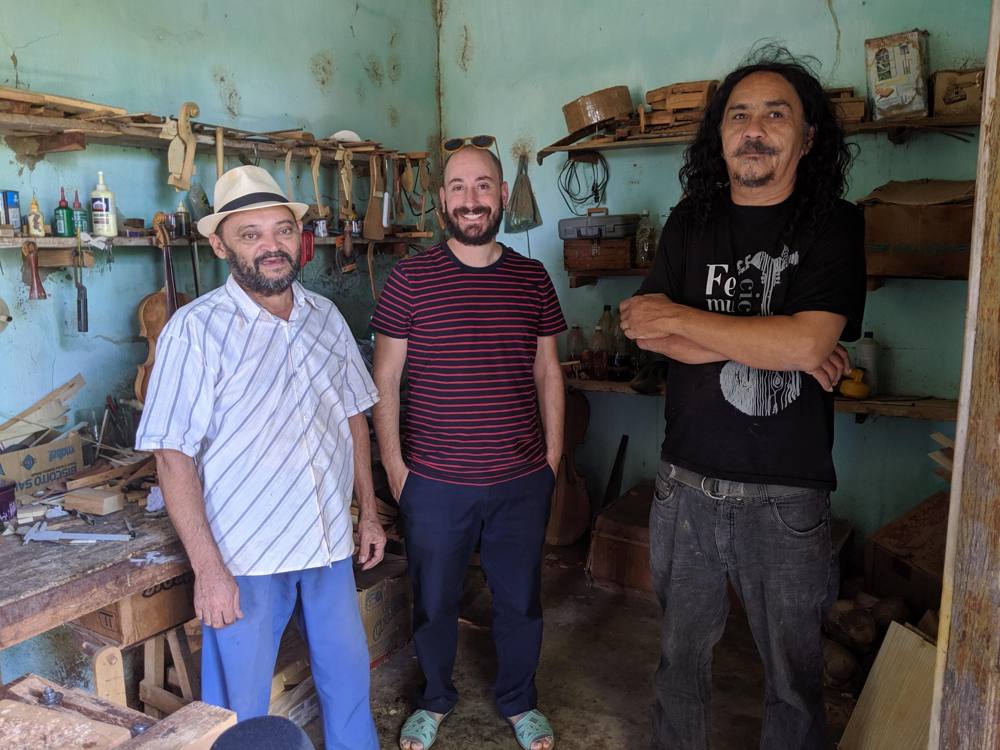 A luthier's workshop in Ceará: the maker in a straw hat, Michael in a striped shirt, and a long-haired colleague among shelves of fiddle parts