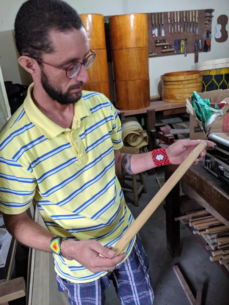A craftsman in a yellow striped shirt holding a long length of pale wood, drum shells stacked behind him