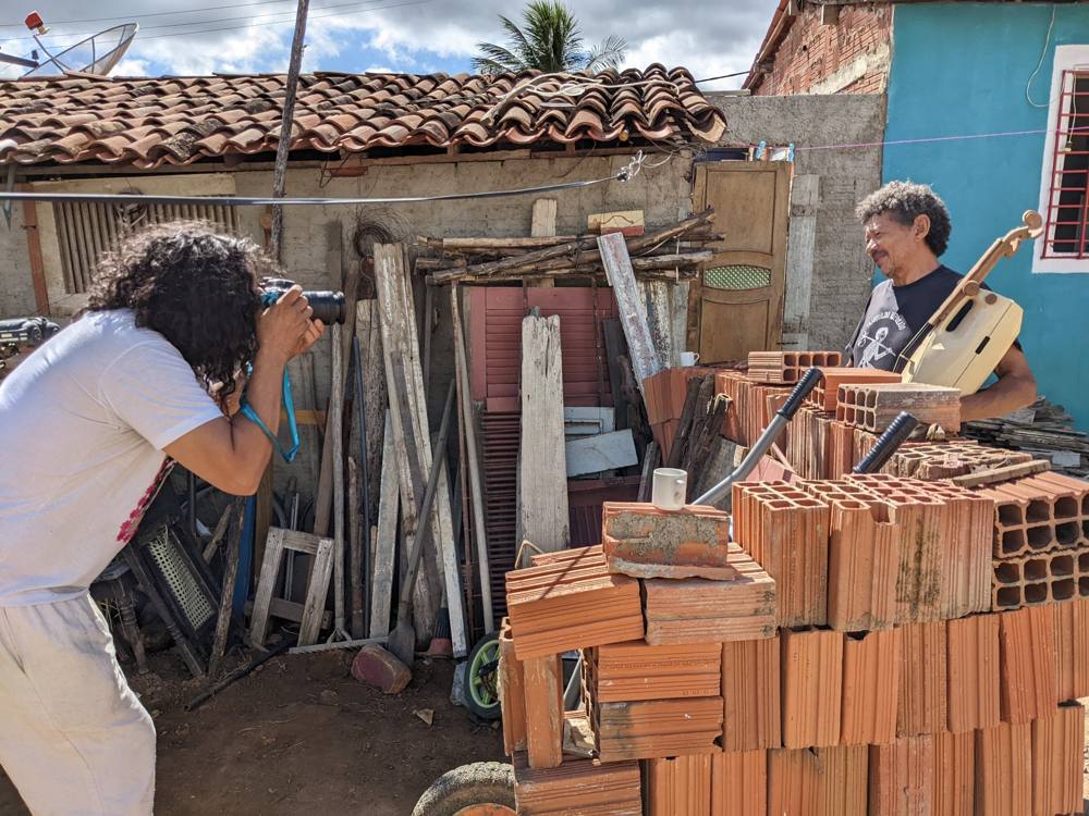 A photographer taking the maker's portrait in his yard as he holds a rabeca made from a plastic gallon jug