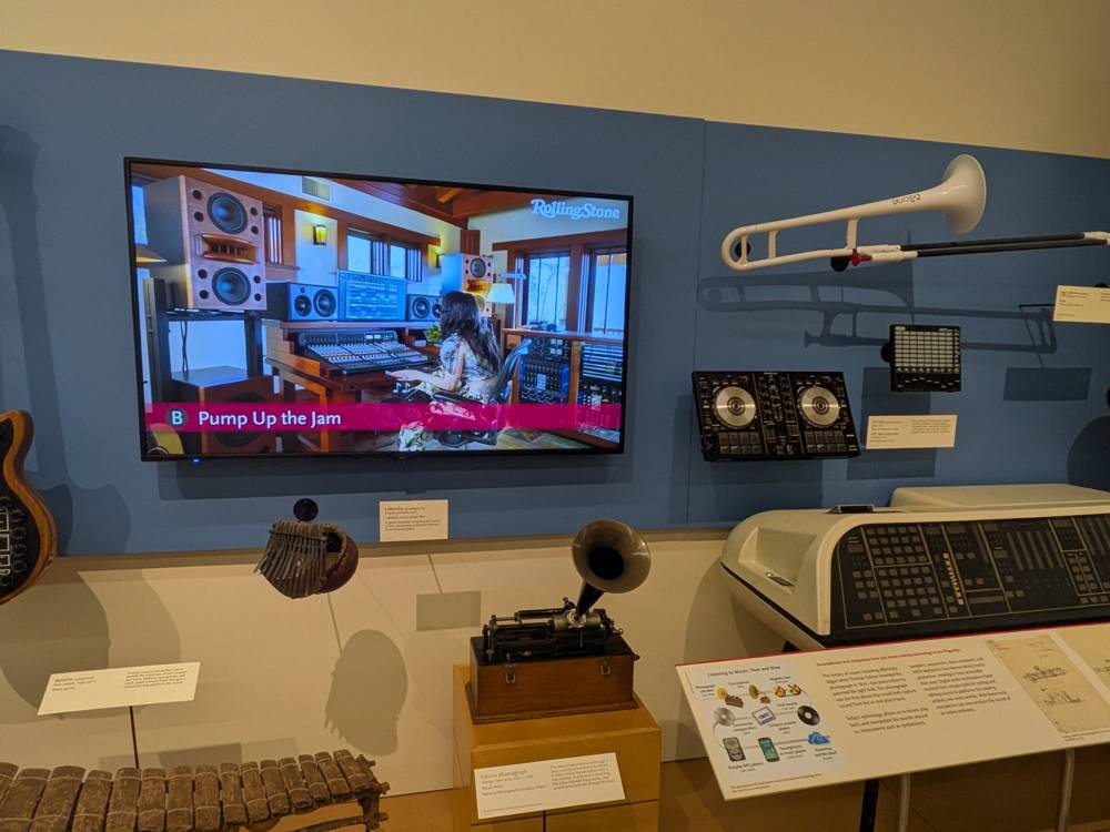 The STEM Gallery's listening wall: Lady Gaga at a studio console on the big screen, a plastic trombone, DJ controllers, and an Edison phonograph below