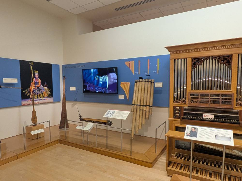 The Mathematical Melodies section: panpipes with ratio graphics, a video screen, the gallery's visible organ, and the mural photograph of Hamid Sidi on the left wall