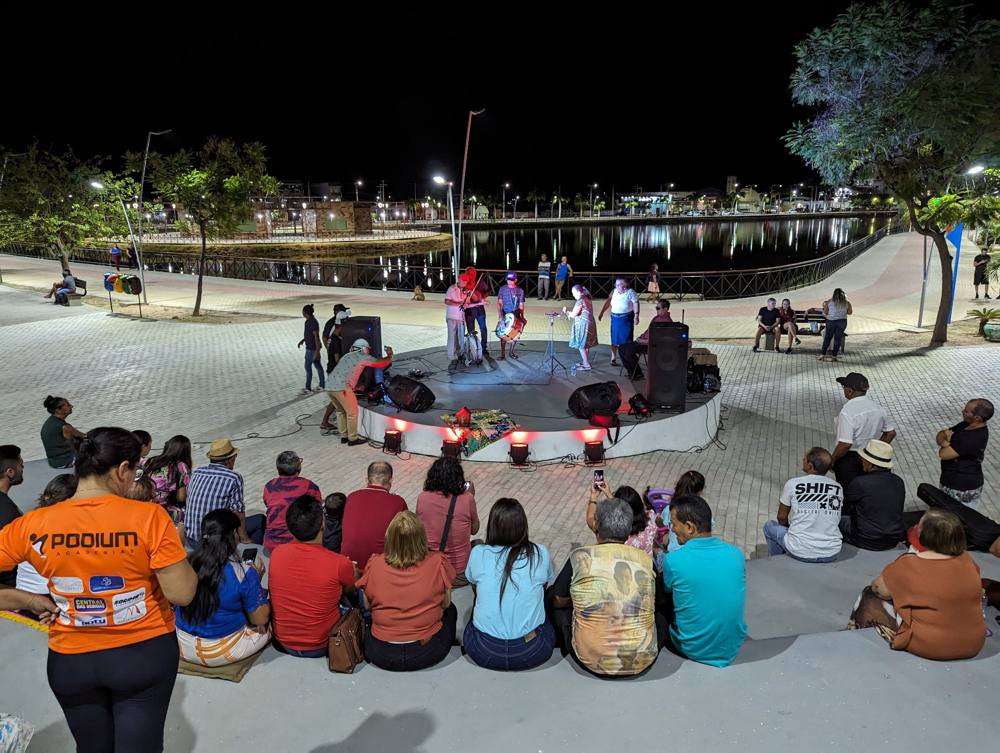 A rabeca trio performing at night on a round lakeside stage before a seated crowd